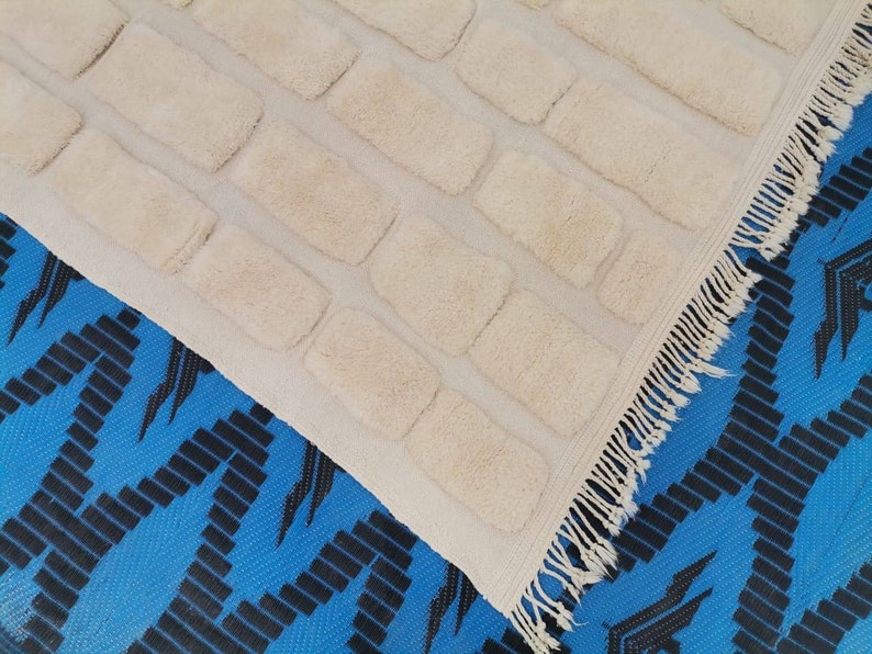 May include: A white rug with a textured brick pattern and a fringe border. The rug is laid on a blue and black patterned surface.