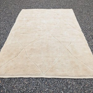 May include: A rectangular, beige rug with a diamond pattern. The rug has a soft, textured surface and is placed on a dark, speckled surface. The rug's edges are finished with a simple border.