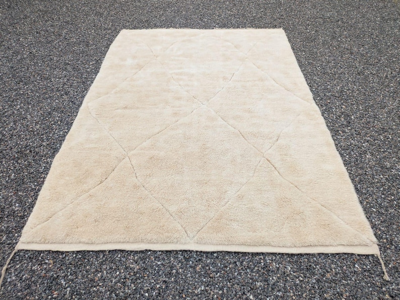 May include: A rectangular, beige rug with a diamond pattern. The rug has a soft, textured surface and is placed on a dark, speckled surface. The rug's edges are finished with a simple border.