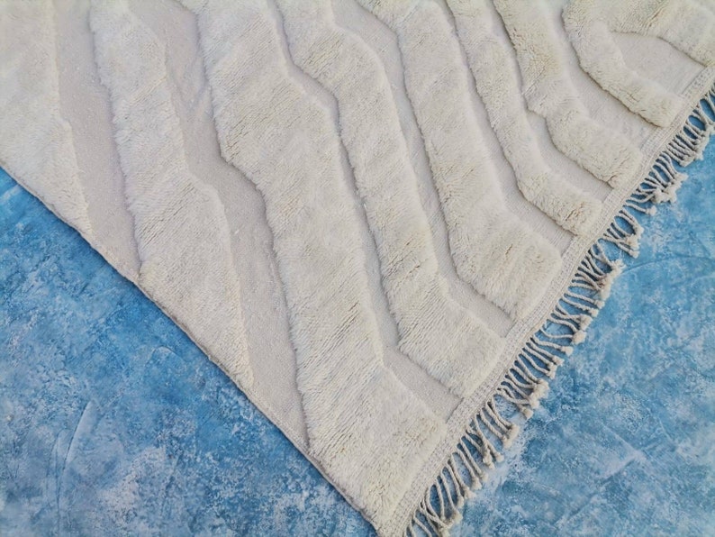 May include: A white, fluffy rug with a chevron pattern and fringe edging. The rug is laid out on a blue surface.