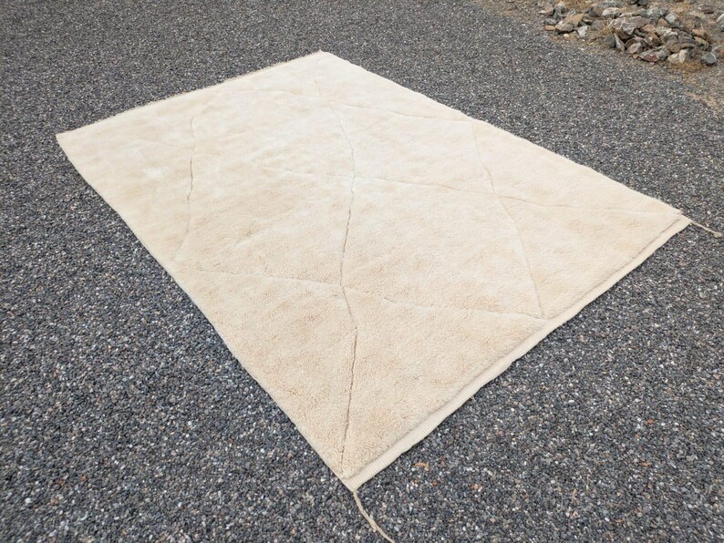 May include: A large, rectangular beige rug with a diamond pattern. The rug is made of a soft, textured material and is laid out on a gravel surface. The rug's edges are finished with a simple border.