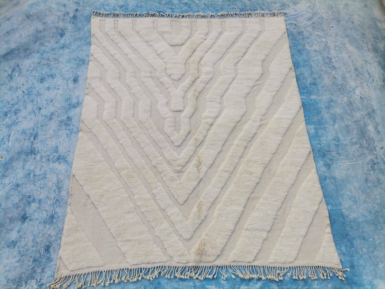 May include: A white, hand-woven rug with a geometric pattern. The rug has a fringe border and is made of natural fibers.
