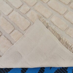 May include: A cream-colored rug with a brick pattern. The rug has a fringe border and is folded in half to show the back side.