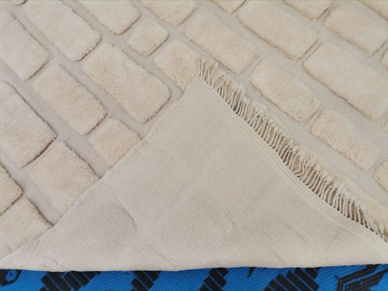 May include: A cream-colored rug with a brick pattern. The rug has a fringe border and is folded in half to show the back side.