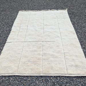 May include: Cream-colored, rectangular rug with a geometric design. The rug features a series of vertical and horizontal lines creating a grid-like pattern. The rug has a soft, textured appearance and a fringe along one edge.