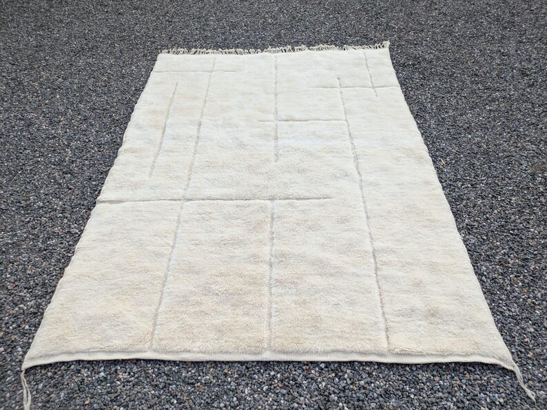 May include: Cream-colored, rectangular rug with a geometric design. The rug features a series of vertical and horizontal lines creating a grid-like pattern. The rug has a soft, textured appearance and a fringe along one edge.