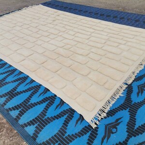 May include: A white rug with a textured brick pattern and fringe edging. The rug is laid out on a blue and black patterned surface.