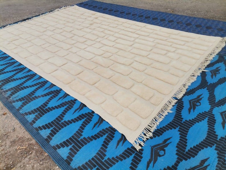 May include: A white rug with a textured brick pattern and fringe edging. The rug is laid out on a blue and black patterned surface.