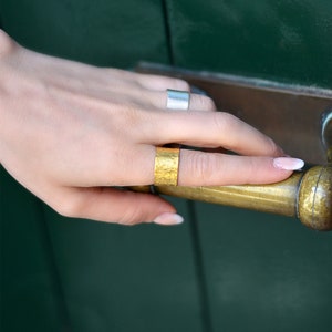 May include: A person's hand wearing two rings. The ring on the index finger is a wide gold band with a hammered texture. The ring on the middle finger is a thin silver band.