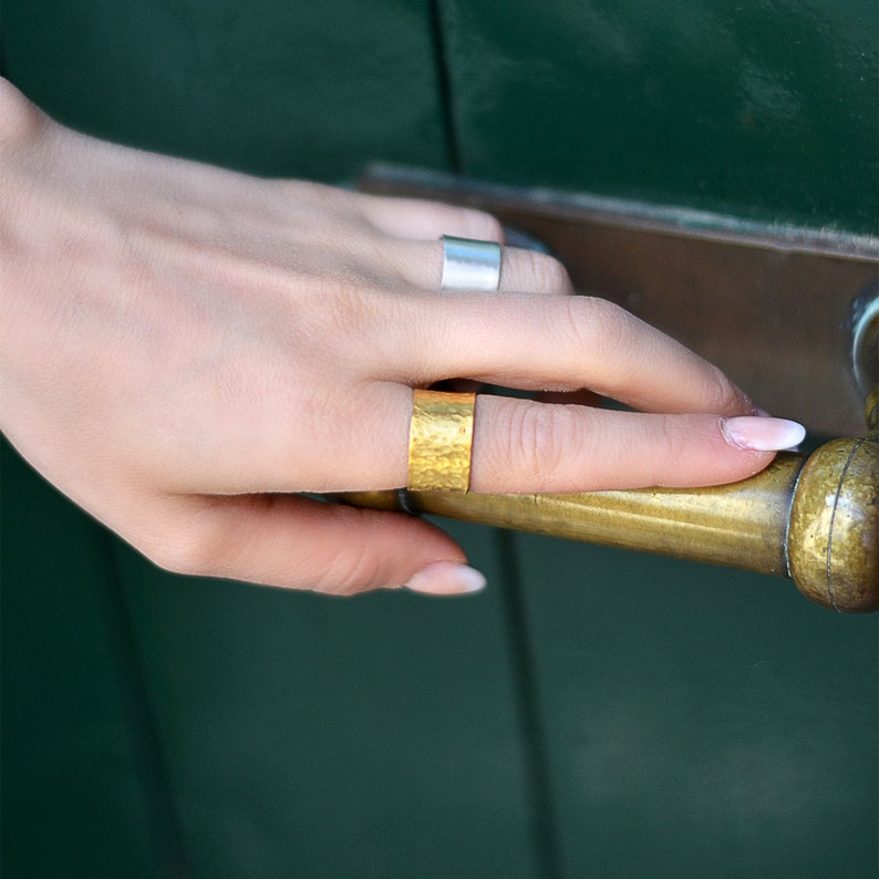 May include: A person's hand wearing two rings. The ring on the index finger is a wide gold band with a hammered texture. The ring on the middle finger is a thin silver band.