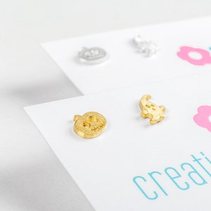 Può includere: Due paia di orecchini dorati su una superficie bianca. Un paio presenta un design a forma di zucca e l'altro paio presenta un design a forma di fantasma. La superficie ha il testo "creating is..." stampato su di essa.