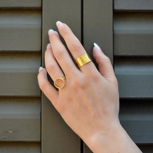 May include: Two gold rings on a hand. One ring is a thin band with a hammered texture. The other ring is a thick circle.