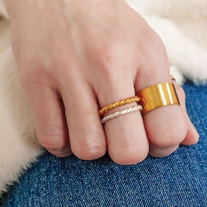 May include: A hand wearing three gold rings. Two of the rings are thin and twisted, and the third is a wider band.