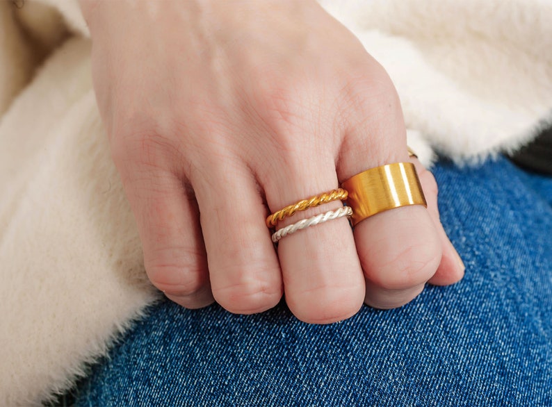 May include: A hand wearing three gold rings. Two of the rings are thin and twisted, and the third is a wider band.