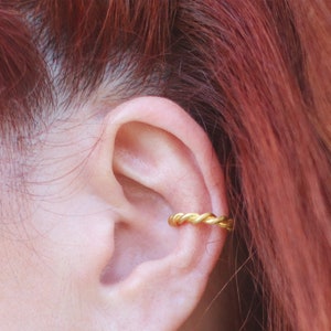 May include: A gold-coloured ear cuff with a braided design. The ear cuff is worn on the upper ear. The background includes red hair and skin.