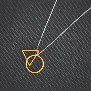 May include: A gold necklace with a circle and triangle pendant. The necklace is on a black fabric background.