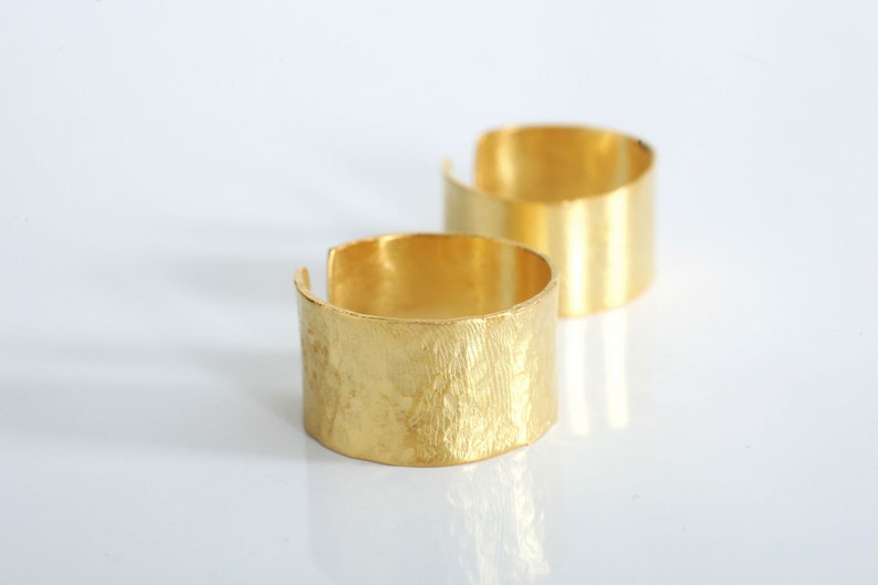 May include: Two gold-toned metal rings. The ring on the left has a textured surface. The ring on the right has a smooth surface.