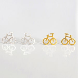May include: A pair of silver and gold bicycle earrings. The earrings are small and delicate, and they feature a simple design of a bicycle.