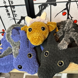 May include: Three crocheted stuffed animals, one brown, one black, and one blue, with large black eyes. The animals are lying on a wooden surface.