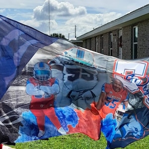 May include: A personalized football flag with a blue, white, and red color scheme. The flag features a football player in a red and white uniform with the number 55 on the front. The flag also features a blue and white football helmet with the number 55 on the side. The flag is waving in the wind.