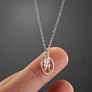 Puede incluir: Un collar de cadena de plata con un colgante que presenta una letra "A" en cursiva dentro de un círculo.