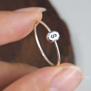 May include: A delicate silver ring featuring a small, octagonal charm with the letter "S" in black. The ring's band is thin and polished, and the charm is centered on the ring. The ring is a piece of jewelry.