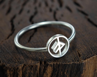 May include: A silver ring with a circular design featuring a rune symbol. The ring is on a wooden surface.