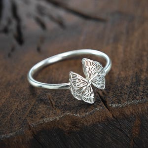 May include: A silver ring featuring a delicate butterfly design. The butterfly is crafted from intricate filigree work and has a slightly open wingspan. The ring is displayed on a dark brown wooden surface.