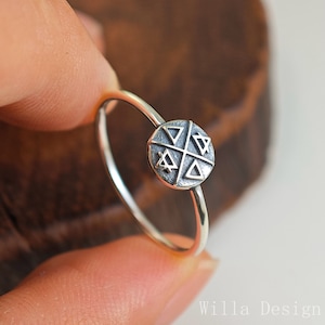 Puede incluir: Un anillo de plata con una pieza central circular y texturizada. La pieza central presenta un diseño geométrico de triángulos y formas de estrella. El anillo está sujeto por los dedos de una persona.