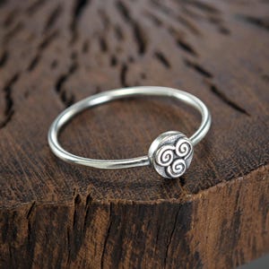 May include: A silver ring with a circular design featuring three stylized spirals. The ring is on a wooden surface.