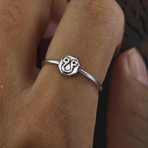 Puede incluir: Un anillo de plata con un diseño hexagonal que presenta un símbolo estilizado. El anillo se lleva en un dedo.