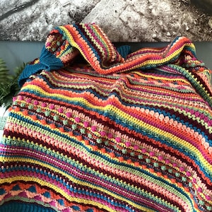 May include: A handmade crochet blanket featuring a vibrant array of horizontal stripes in shades of teal, yellow, orange, pink, and deep red. The textile displays a variety of intricate stitch patterns, including rows of raised bobbles and textured clusters.