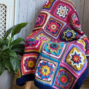 Könnte beinhalten: Eine bunte gehäkelte Wolldecke mit einem Blumenmuster in den Farben Rosa, Lila, Grün, Gelb und Blau. Die Decke liegt über einem Holzstuhl.