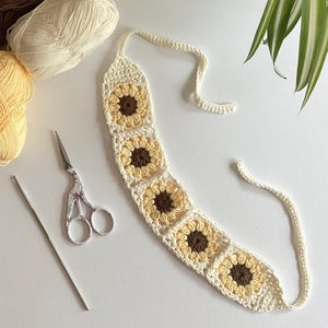 May include: A white crocheted sunflower garland with brown centres. The garland is made of multiple crocheted squares with a sunflower design. The garland is tied together with a white string.