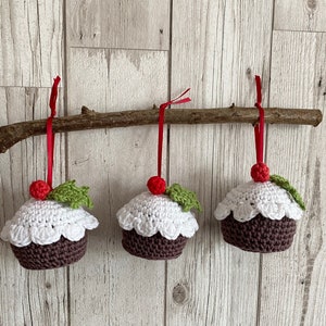 May include: Three crocheted Christmas ornaments shaped like pudding with white icing and red holly berries. The ornaments are hanging from a branch with red ribbon.