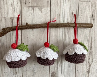 May include: Three crocheted Christmas ornaments shaped like pudding with white icing and red holly berries. The ornaments are hanging from a branch with red ribbon.