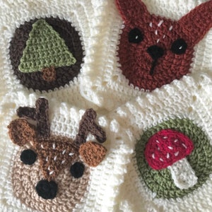 Könnte beinhalten: Eine weiße Häkeldecke mit verschiedenen Tier- und Naturmotiven. Die Motive umfassen ein Reh, einen Pilz, einen Baum und einen Fuchs. Die Decke ist aus weichem, flauschigem Garn gefertigt.