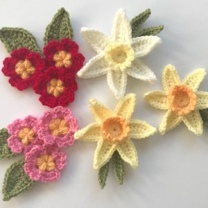 Op de afbeelding: Vijf gehaakte bloemenversieringen met groene bladeren. De bloemen zijn in tinten rood, roze, geel en wit.