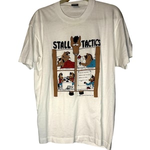 May include: White t-shirt featuring a cartoon horse with the words "STALL TACTICS" above a four-panel comic strip. The comic strip depicts horses in various scenarios. The shirt is a crew neck with short sleeves.