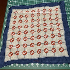 May include: A white silk scarf with a blue border and a repeating pattern of red interlocking Gs. The scarf has the word "GUCCI" printed in the bottom right corner.