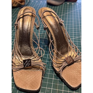 Può includere: Un paio di sandali in pelle beige con tacco alto, dettagli dorati e il logo Louis Vuitton sulle fasce.