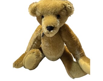 Puede incluir: Un oso de peluche vintage marrón con ojos negros y una nariz de botón. El oso está sentado con las piernas extendidas.