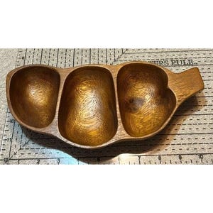 May include: A wooden serving dish with three connected compartments and a handle. The dish is made of a warm-toned wood, showcasing the natural grain. The compartments are oval-shaped, perfect for serving snacks or dips.