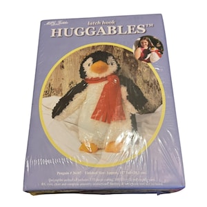 Peut inclure: Un kit de crochet en forme de pingouin. Le pingouin, avec une écharpe rouge, est placé sur un cercle jaune sur un fond violet clair. Le kit comprend une toile, de la laine, du feutre et des instructions. La taille finie est d'environ 38,1 cm de haut.