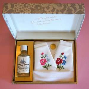 May include: A white gift box with a gold trim and a gold interior. The box contains two white handkerchiefs with embroidered flowers and a small bottle of clear liquid labeled "Echt Linloch Wasser". The box lid has the text "Allerfeinste Handmaschinen Stickerei" printed on it.