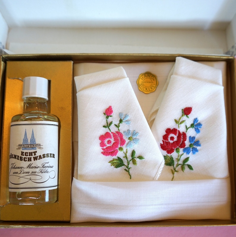 May include: A gift box containing two white handkerchiefs with embroidered pink and red roses and blue flowers. The box also contains a clear glass bottle with a white label that reads "Echt M&uuml;nich Wasser" and "Johann Maria Farina am Dom zu K&ouml;ln".