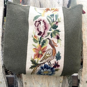 Könnte beinhalten: Eine grüne Stofftasche mit einem cremefarbenen Einsatz, der mit einem gestickten Blumendesign mit einem Vogel verziert ist. Die Stickerei ist in den Farben Rosa, Orange, Grün, Braun und Blau gehalten.