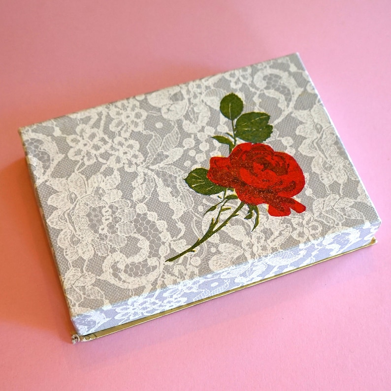 May include: A white box with a lace pattern and a red rose printed on the lid. The box is closed.