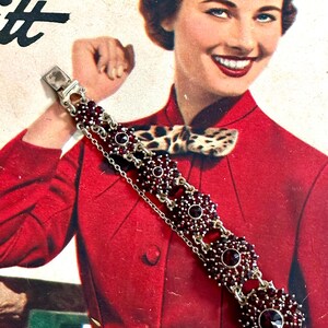 Puede incluir: Un brazalete de plata con una cadena y varios racimos de piedras de granate rojo. El brazalete está sobre un fondo rojo.