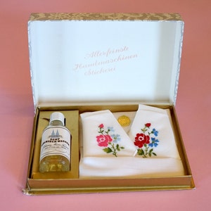May include: A white box with a gold trim and a white lid with the text "Allerfeinste Handmaschinenstickerei" in pink lettering. The box contains two white handkerchiefs with embroidered flowers and a small clear bottle with a label that reads "Echt Almrich Wasser".
